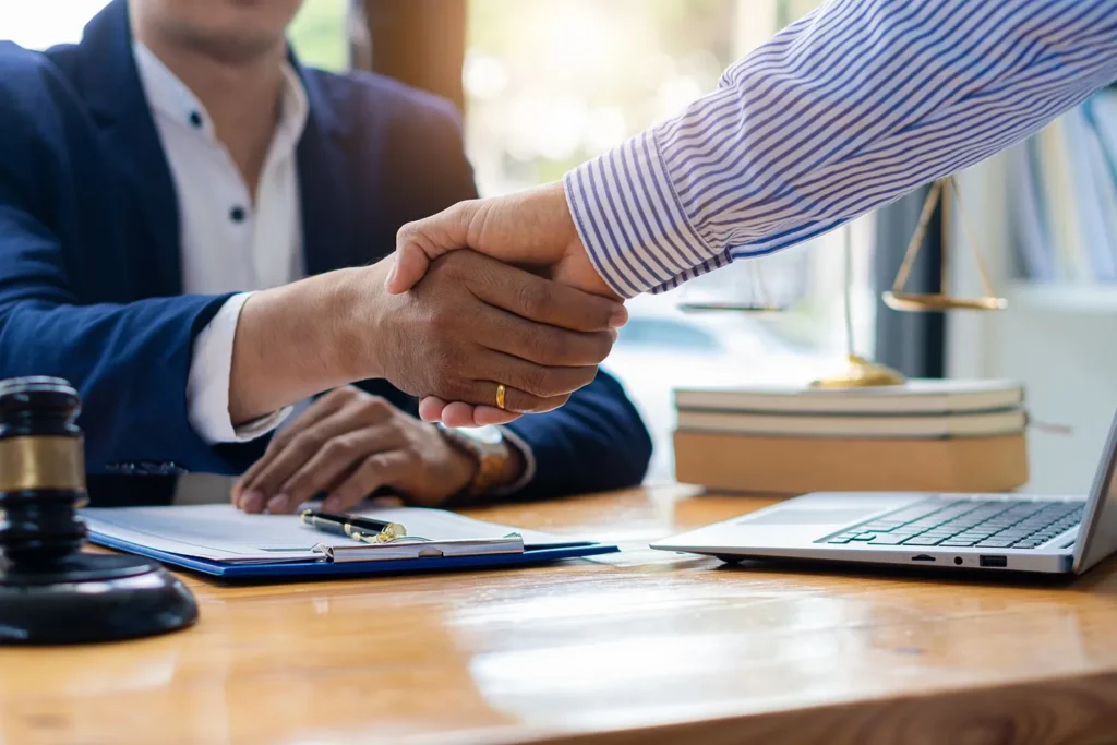 Two men in professional clothes shaking hands, there is a legal gavel, clipboard and laptop on the desk.